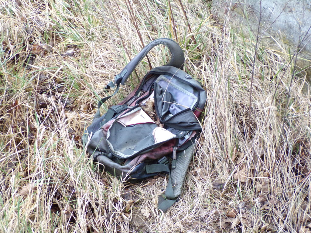 Abgenutzter Rucksack mit offenen Fächern auf dem Boden in hohem Gras.