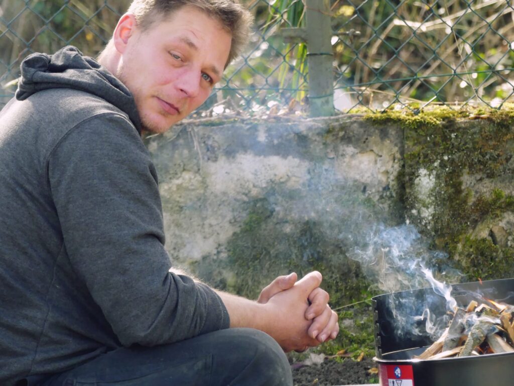 Mann in grauem Sweatshirt sitzt neben einem kleinen Feuer mit aufsteigendem Rauch.