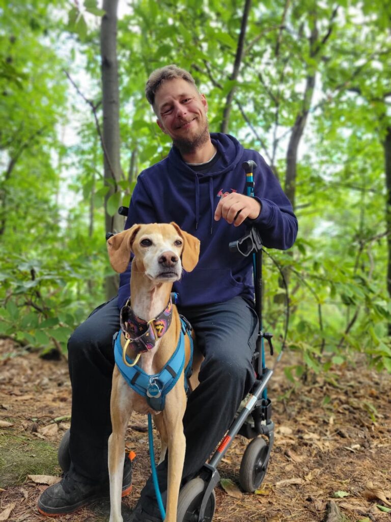 Ein Mann sitzt auf einem Rollator und hat einen Hund an der Leine. Beide sind im Freien.
